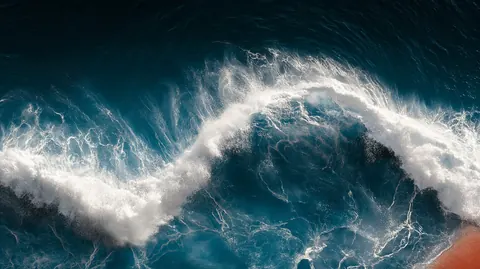 A white crest of foam curls across a deep blue-green sea, seen from above like a wave caught mid-turn.